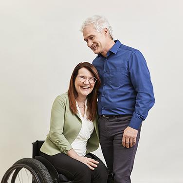 Mario stands next to Sylvie in her wheelchair, they are embracing.