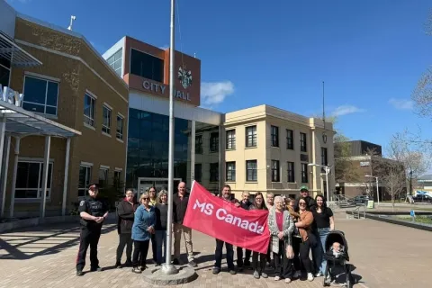 A group of people stands outside a city hall building on a sunny day, gathered around a flagpole while holding a red MS Canada banner. 