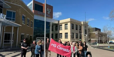A group of people stands outside a city hall building on a sunny day, gathered around a flagpole while holding a red MS Canada banner. 