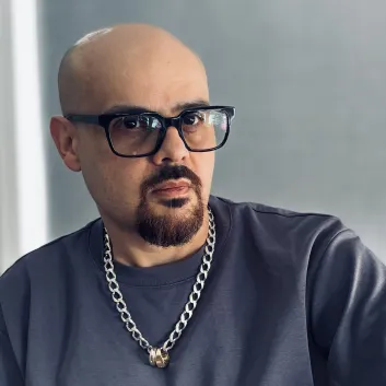 Miguel in gray t-shirt and chain necklace, wearing glasses, seated indoors.